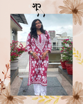A woman stands on a rooftop terrace wearing a maroon pink and white printed kurta with white pants. The photo is framed with a decorative beige border featuring floral illustrations and a black symbol at the top.