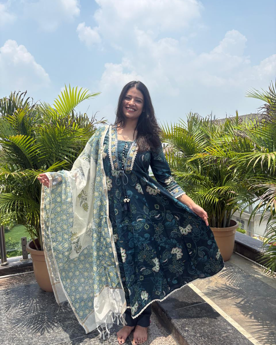 A woman stands outdoors on a rooftop terrace wearing a dark teal blue printed Anarkali kurta with a matching dupatta. The kurta features intricate floral patterns and gold embroidery on the neckline.