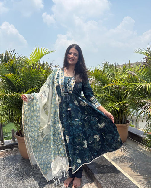 A woman stands outdoors on a rooftop terrace wearing a dark teal blue printed Anarkali kurta with a matching dupatta. The kurta features intricate floral patterns and gold embroidery on the neckline.